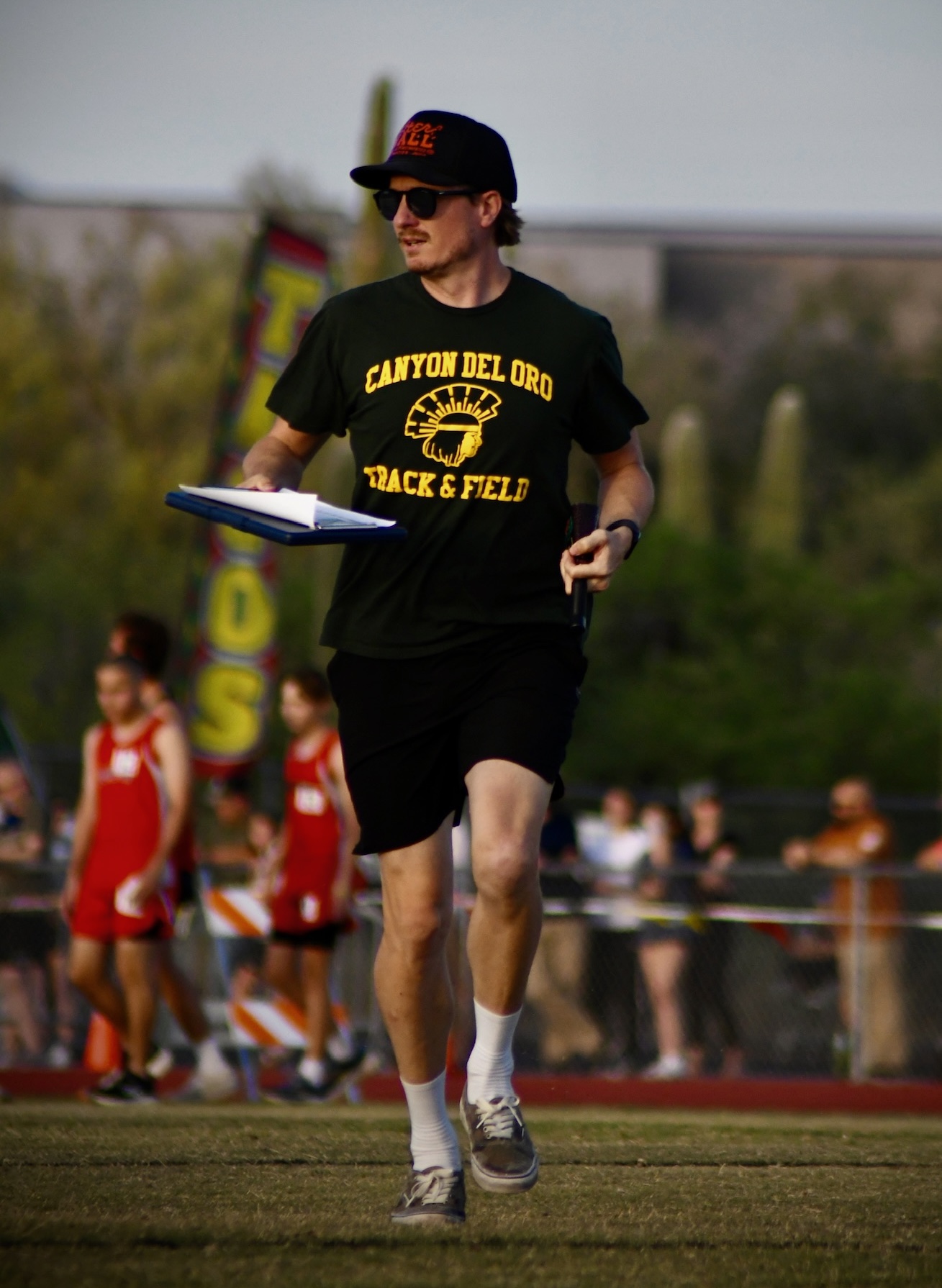 Coach Hunter Babler at the track, holding his coaching clipboard and wearing his Canyon del Oro Track & Field shirt.